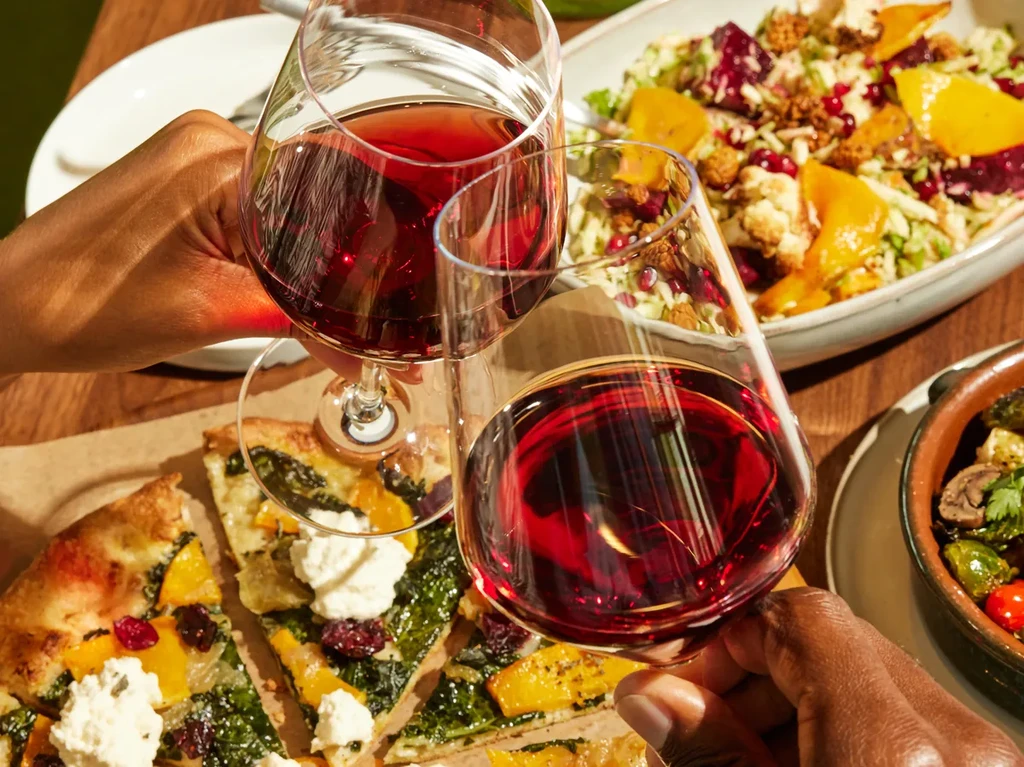 Top-down view of hands clinking red wine glasses above food.