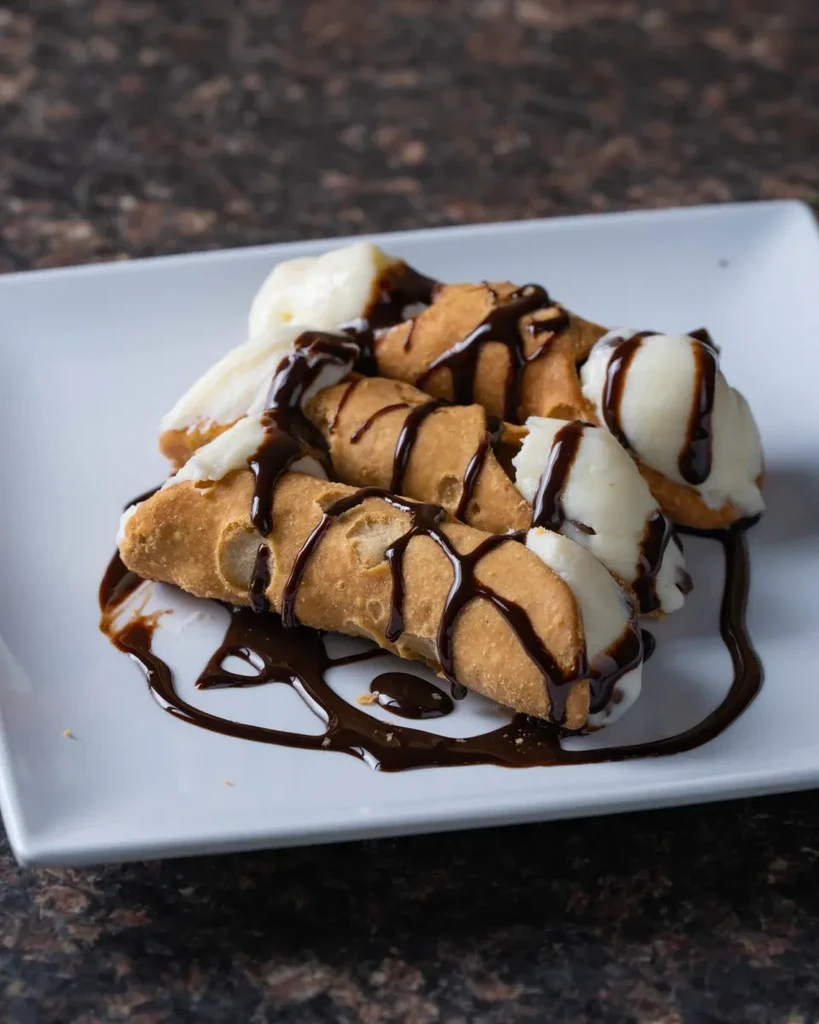 Three crispy cannoli with white filling and chocolate drizzle on a plate.