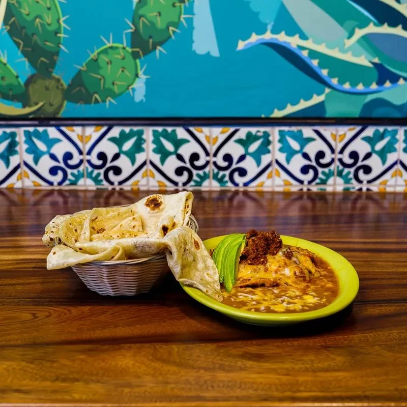 Close-up of enchiladas with avocado, tortillas, and a Mexican mural.