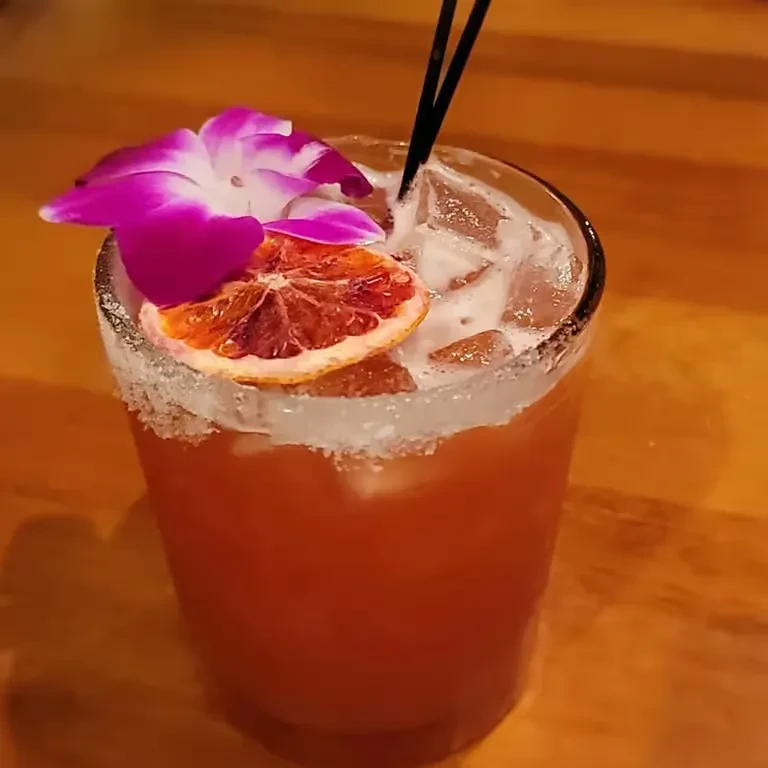 Reddish-orange cocktail with orchid, dried orange, and straws on wood.