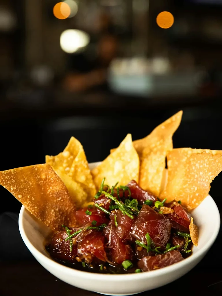 Tuna poke in white bowl with wonton chips and green herbs.