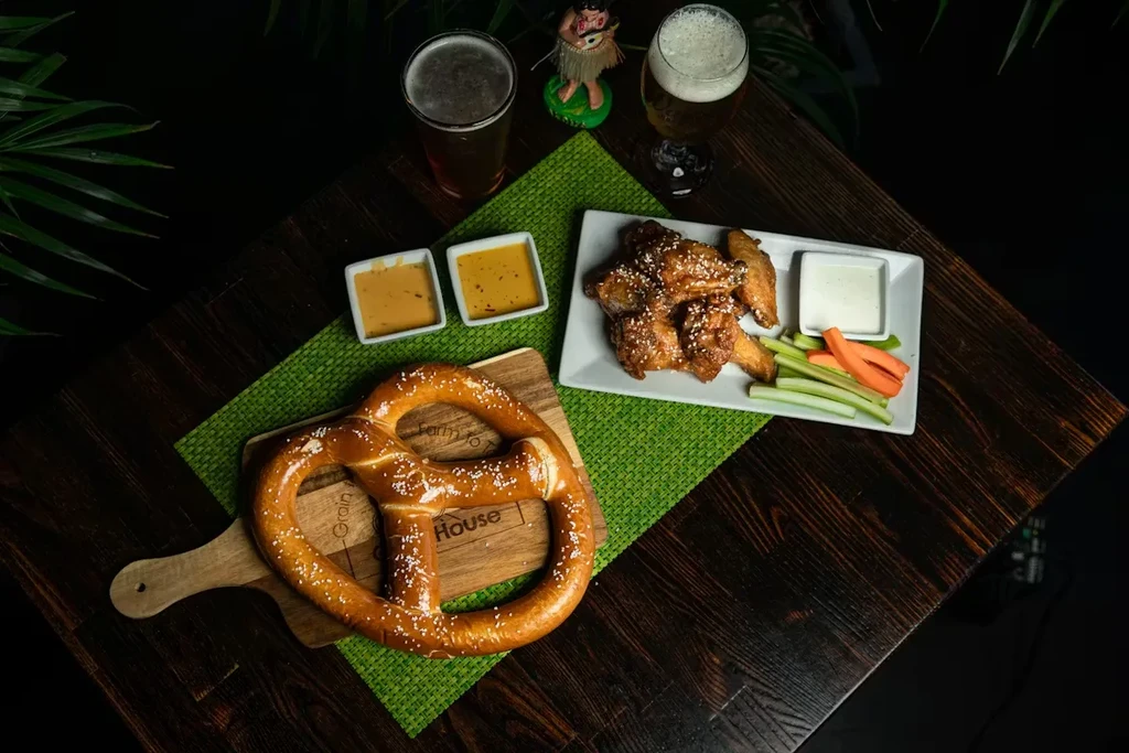Top-down view of a soft pretzel, chicken wings, sauces, and two beers.