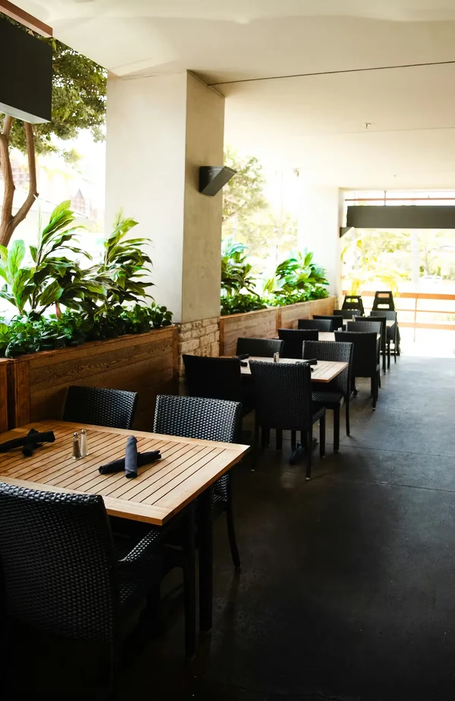Covered restaurant patio with empty wooden tables, dark chairs, and green plants.