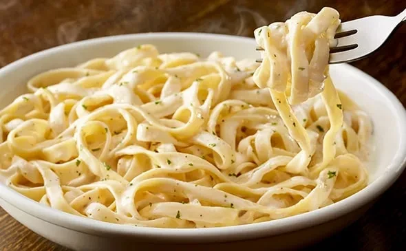 Fork lifts steaming creamy fettuccine Alfredo with herbs from a white bowl.