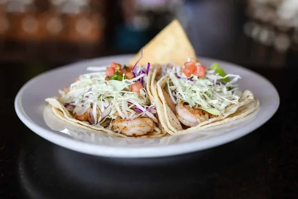 Two shrimp tacos with cabbage, pico de gallo, and a chip on a plate.