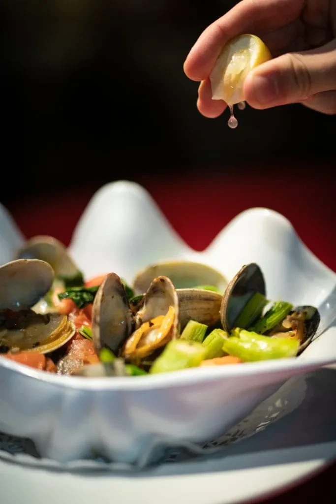 Hand squeezing lemon over clams and green vegetables in a white bowl.