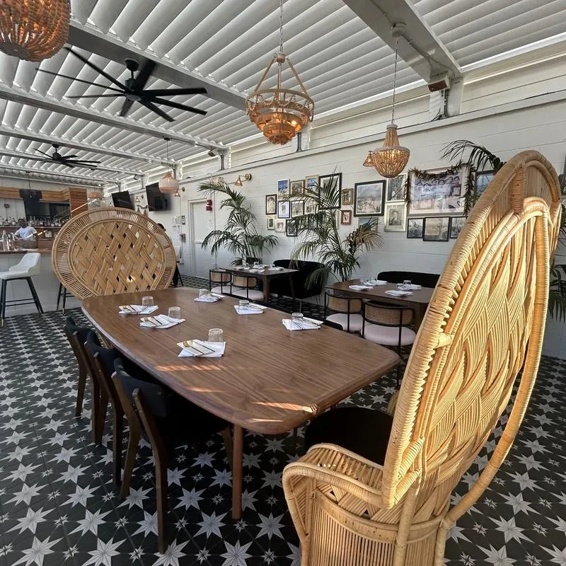 Restaurant patio with wooden table, rattan chairs, plants, and patterned floor.