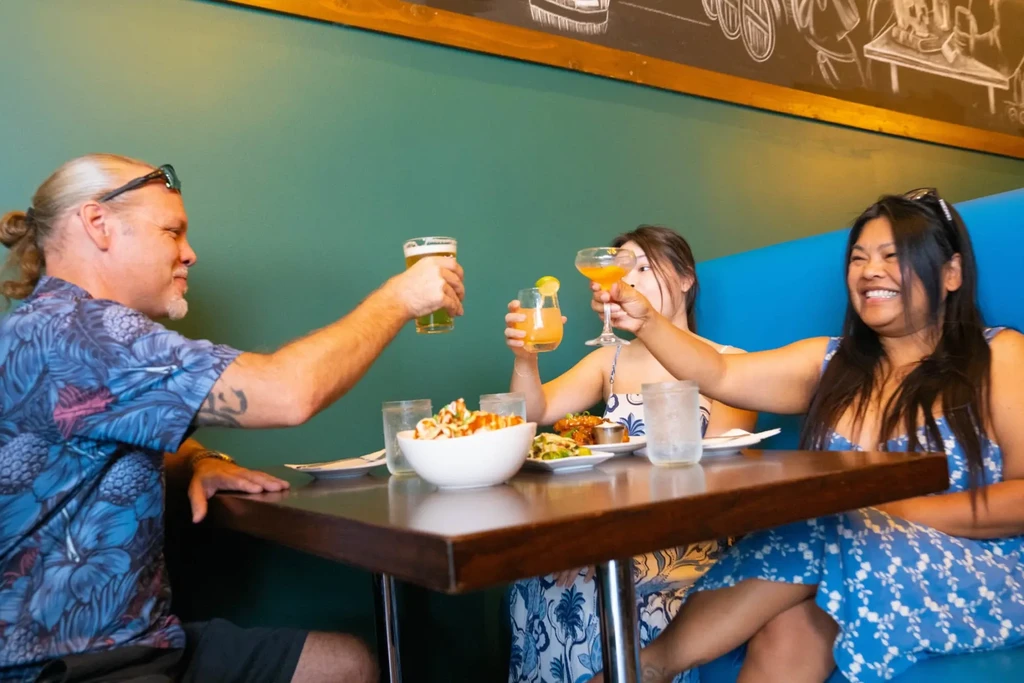 Three people toast with drinks and food in a vibrant restaurant.