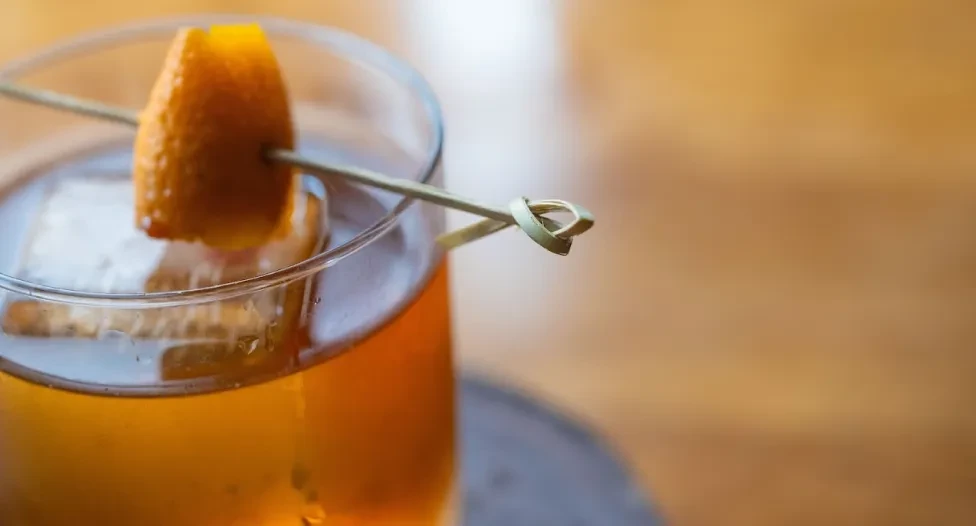 Amber cocktail with large square ice and orange peel garnish.