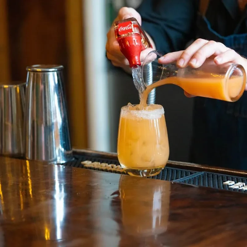 Hands pour from carafe and miniature Coca-Cola bottle into a rimmed glass.