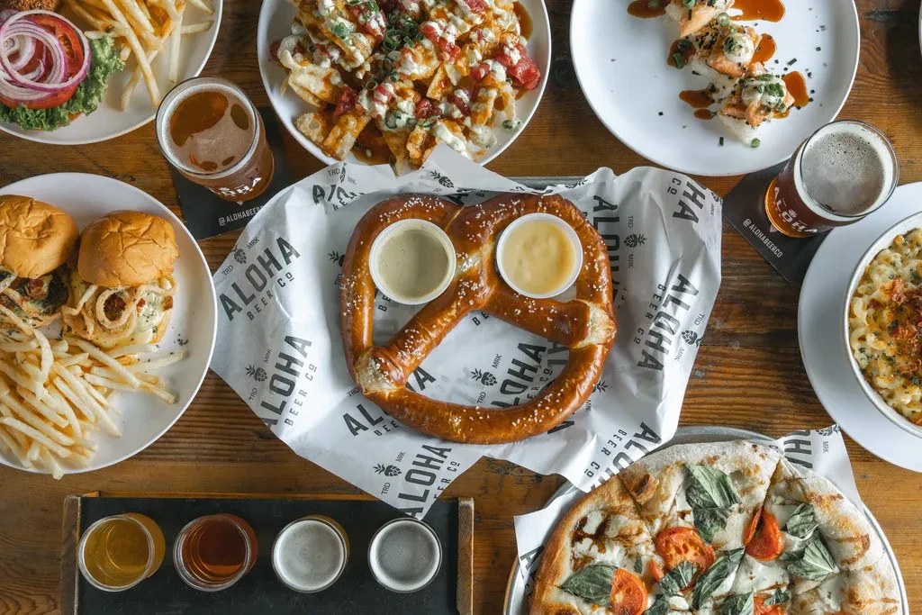 Overhead view of a wooden table with pretzel, sushi, pizza, and Aloha Beer.