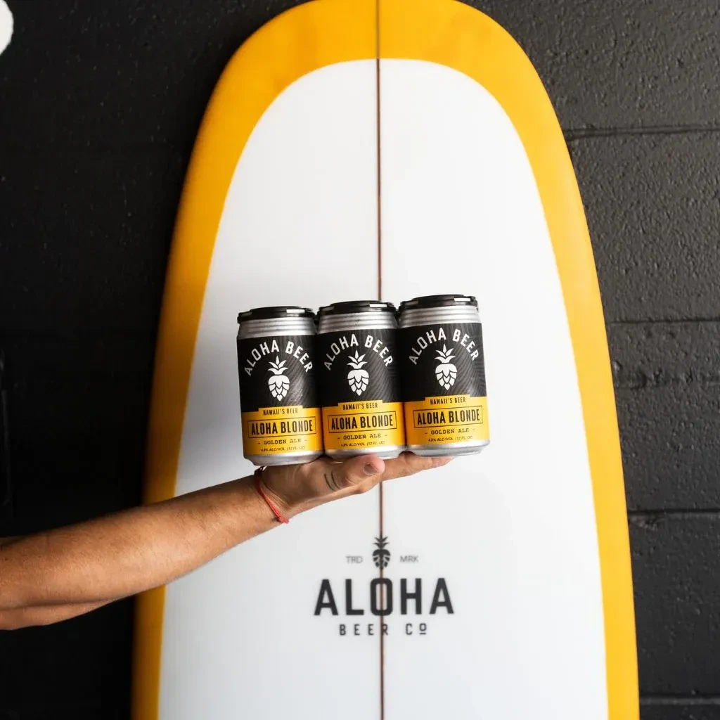 Hand holding three Aloha Blonde beer cans; surfboard behind.