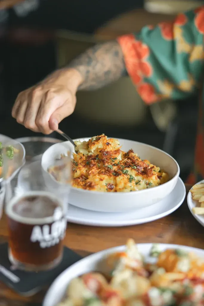 Tattooed hand scooping baked mac and cheese with a fork from a bowl.