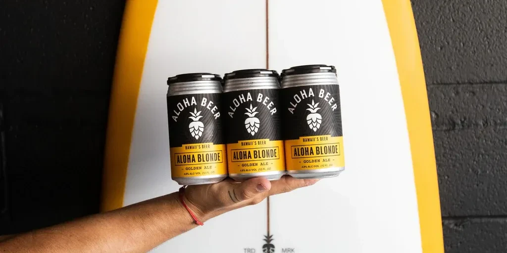 A hand holds three yellow and black Aloha Blonde beer cans.