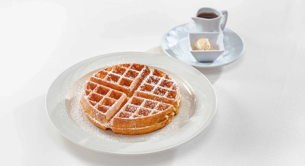 Golden-brown waffle with powdered sugar, syrup, and butter on white dishes.