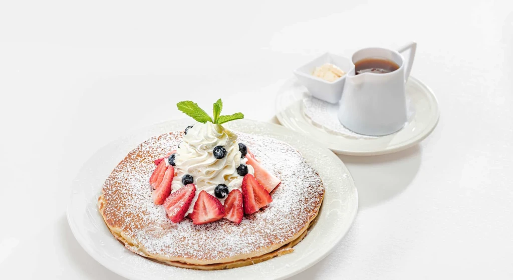 Golden pancakes with powdered sugar, whipped cream, berries, and syrup.