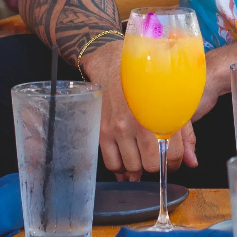 Two drinks, orange cocktail with flower, by a tattooed arm on table.
