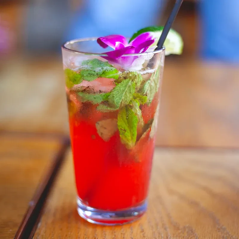 Red cocktail with mint, lime, and magenta orchid on wood.