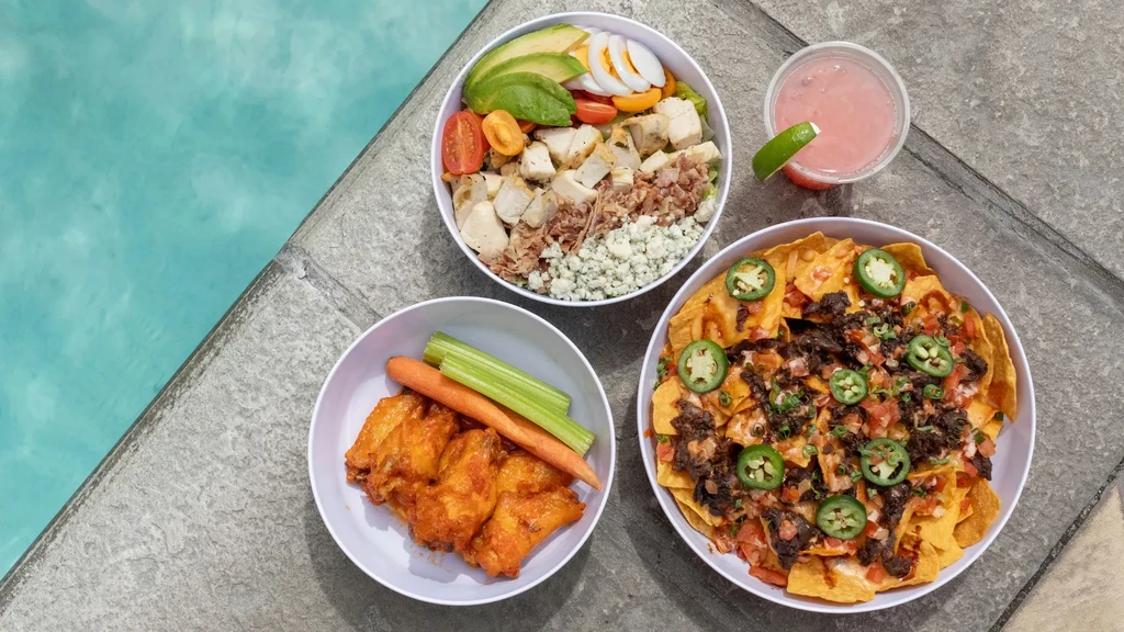 Overhead: Cobb salad, nachos, wings, and pink drink by a pool.
