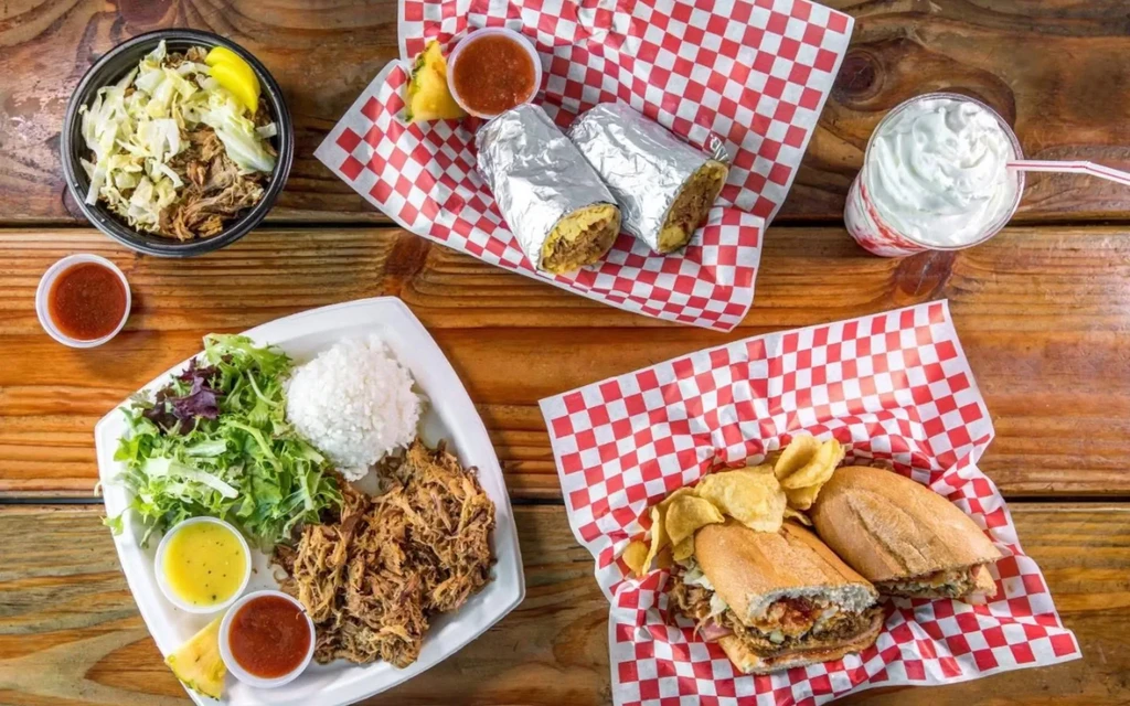 Rustic wooden table filled with burritos, meat, salad, sandwich, and drink.