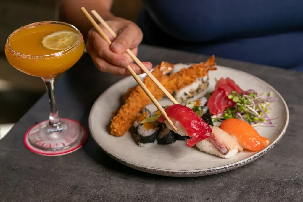 Chopsticks lift tuna nigiri from sushi, sashimi, tempura, and a cocktail.
