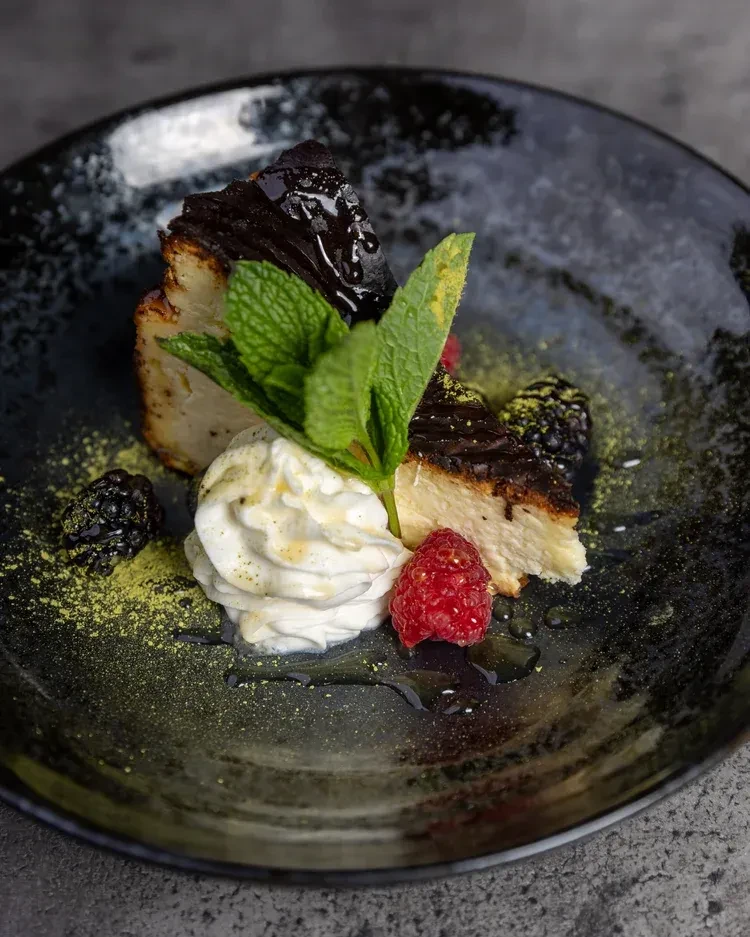 Glazed cheesecake slice with berries, mint, whipped cream, and green powder.