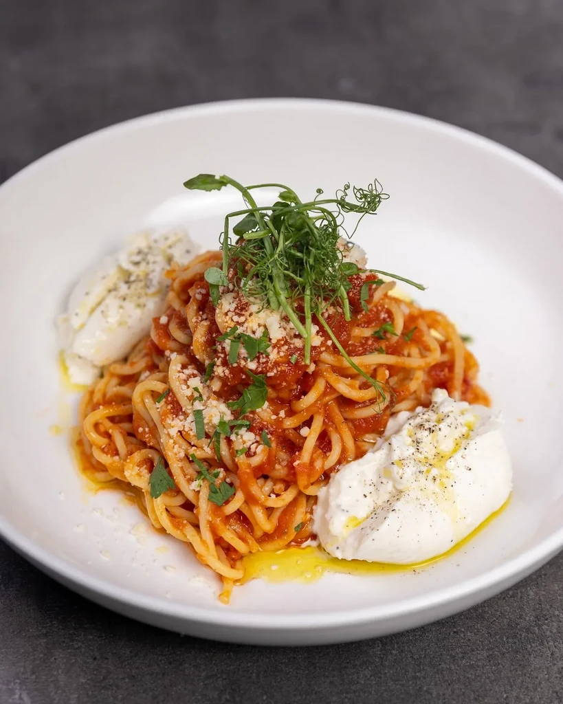 Spaghetti with tomato sauce, Parmesan, microgreens, and burrata in a white bowl.