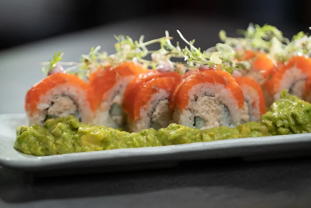 Plated salmon sushi rolls garnished with microgreens and mashed avocado.