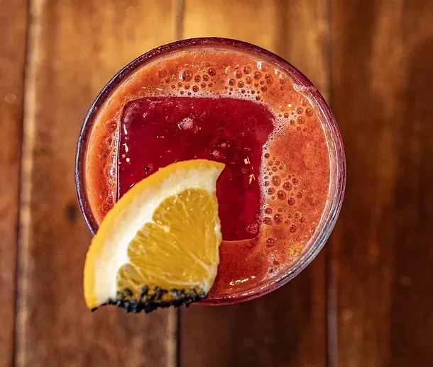 Overhead: Frothy reddish-orange drink, square ice, orange slice on wood.