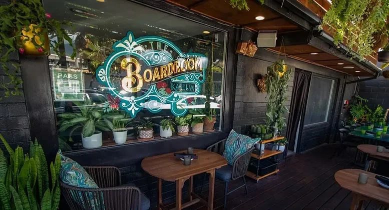 "THE BOARDROOM KALIUA" exterior with ornate illuminated sign and lush outdoor seating.