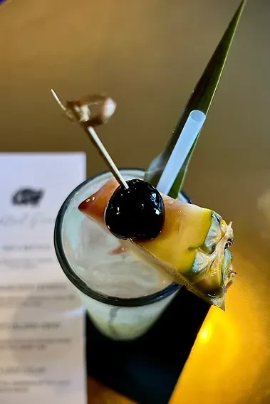 Light-colored cocktail with pineapple wedge, cherry, green leaf, and straw.