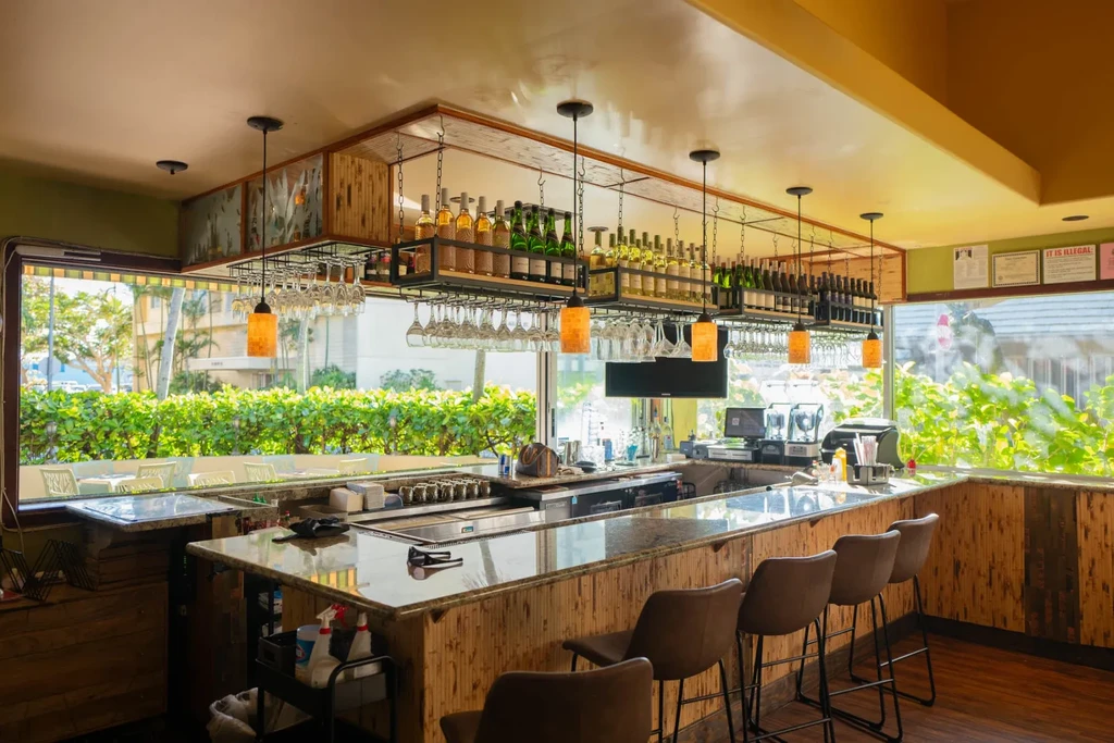 Modern bar with stools, shelves of bottles, and tropical outdoor view.