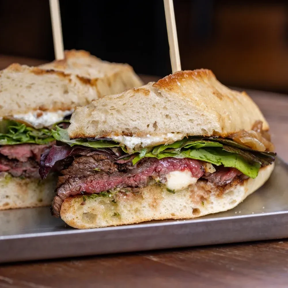 Halved artisan steak sandwich with lettuce, onions, and green pesto.