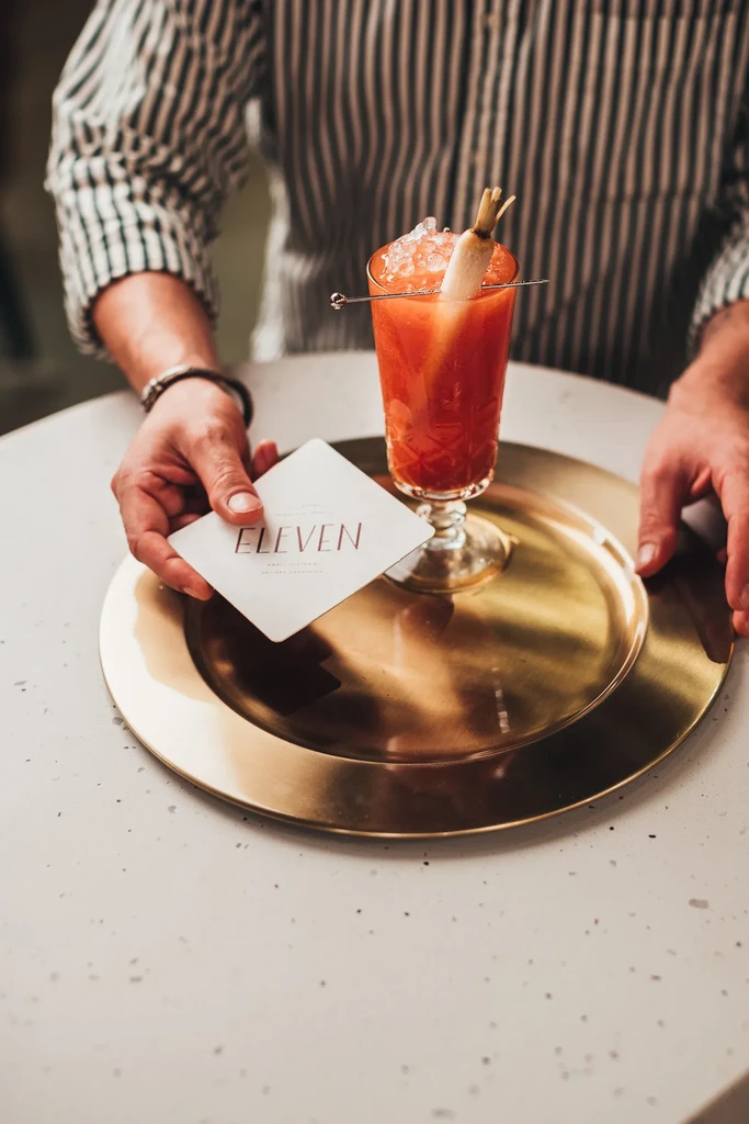 Red cocktail with white carrot garnish, hand holding 'ELEVEN' coaster.