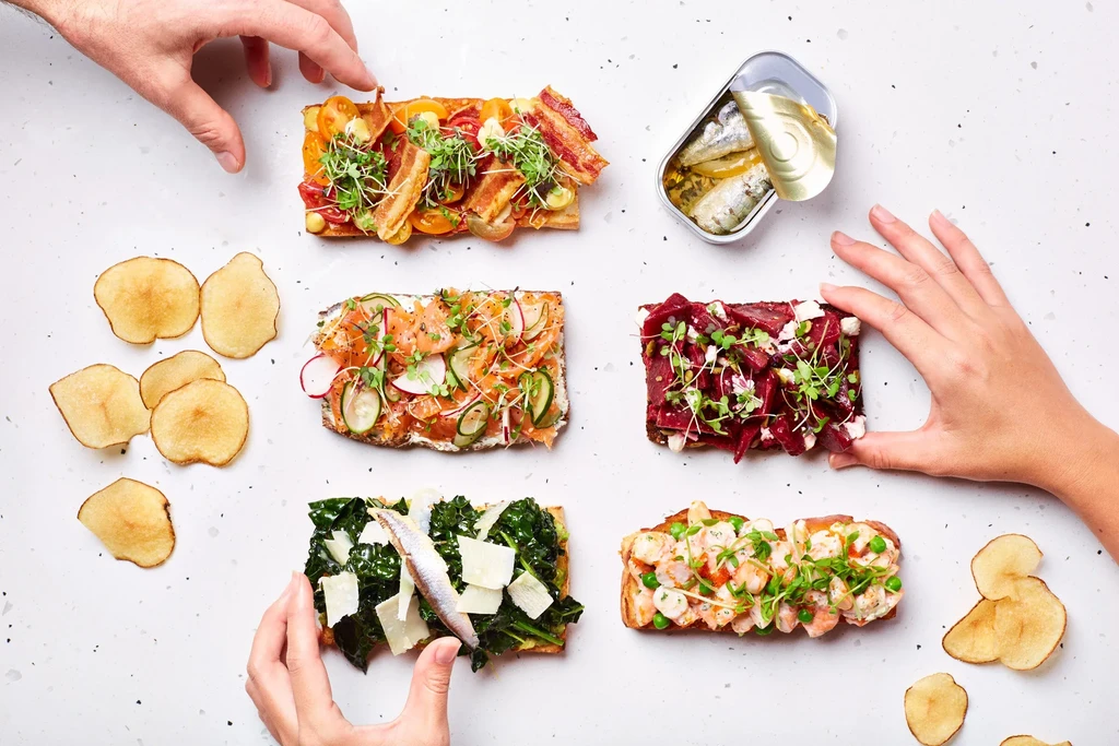 Five open-faced sandwiches with diverse toppings, a sardine tin, and hands.