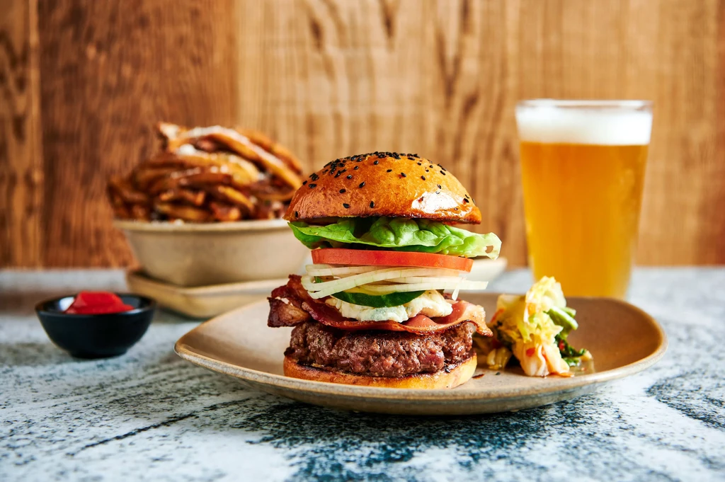 Gourmet burger, kimchi, fries, beer, and red sauce on table.