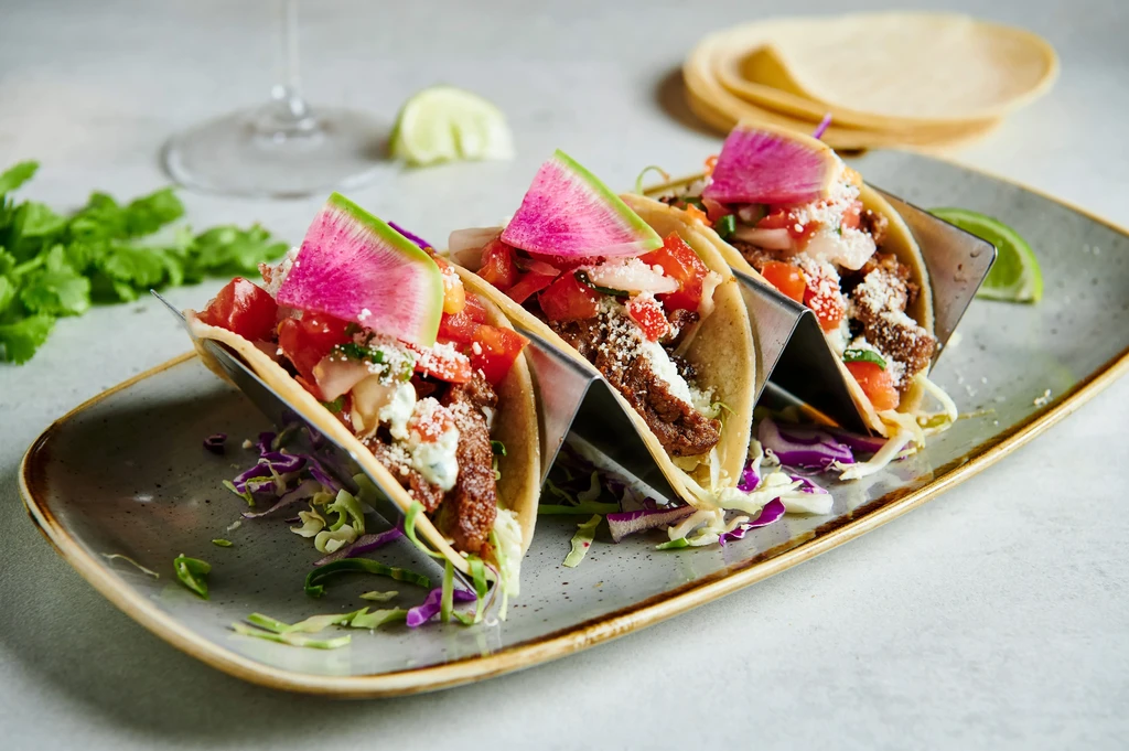 Three tacos with grilled meat, cheese, radish, and green sauce.