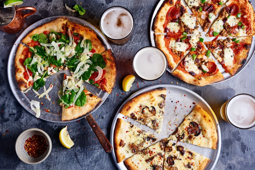 Overhead shot: Three distinct pizzas, beers, and a cocktail on a dark table.