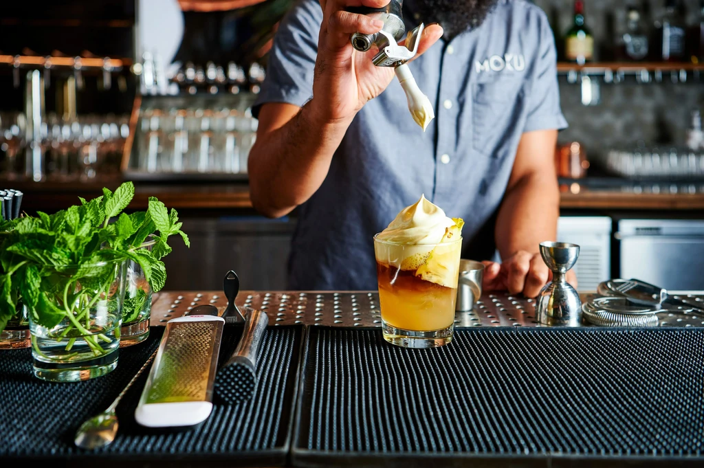 Bartender adds foam to a layered cocktail with pineapple garnish.