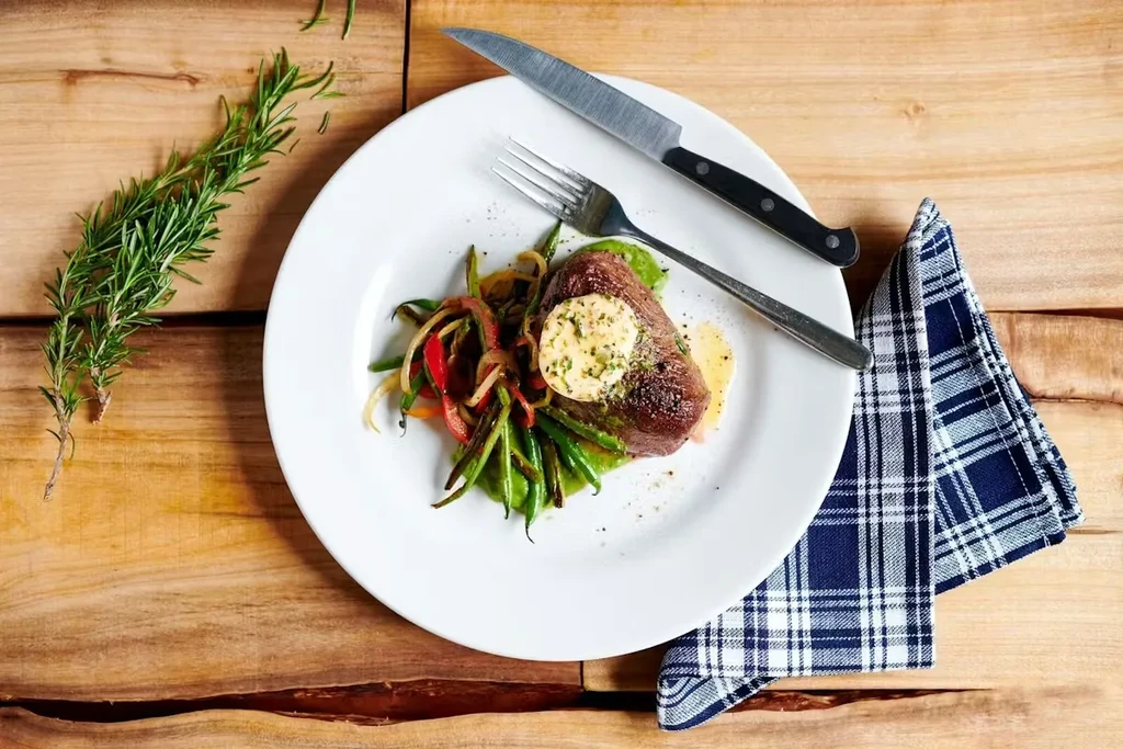 Top-down: Medium-rare steak with herb butter, vegetables, cutlery, wooden table.