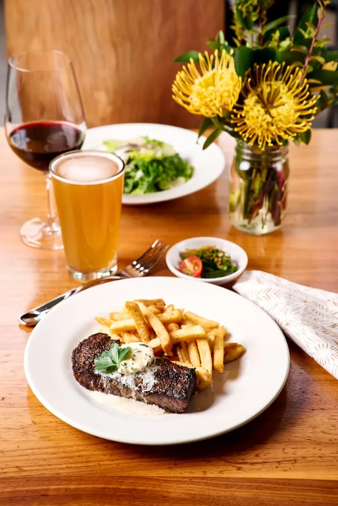 Steak, fries, wine, beer, salad, and flowers on a dining table.