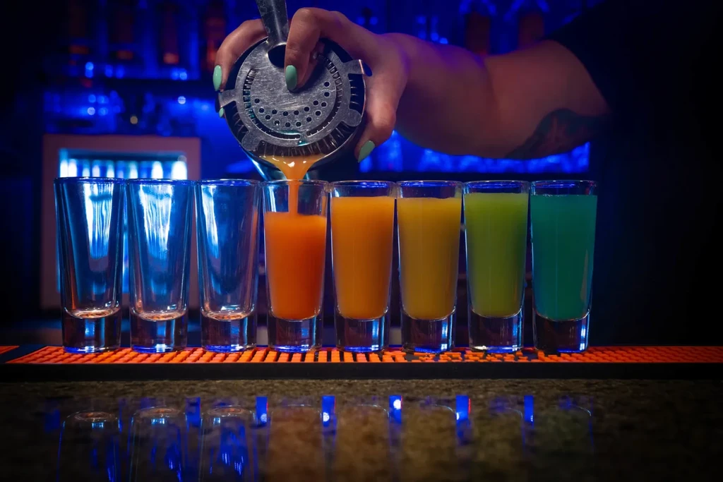 Bartender pours colorful shots at a dimly lit bar with blue light.