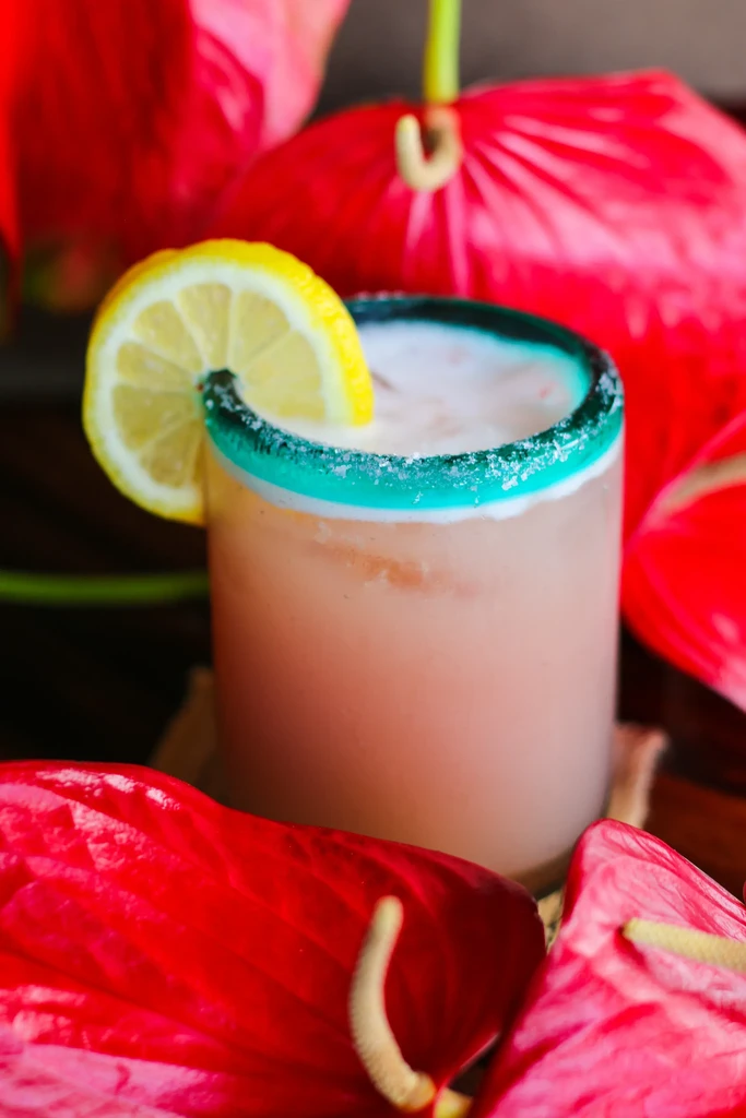 Peachy-pink cocktail with teal sugar rim, lemon slice, and anthurium flowers.