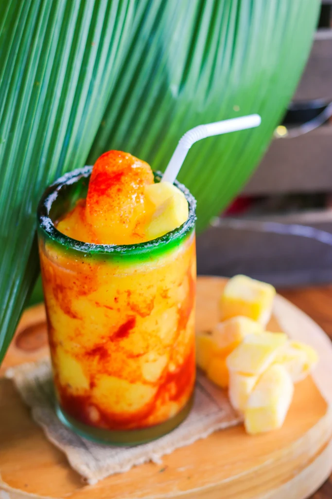 Vibrant mango slushie with chamoy, garnished, on wood, tropical leaf background.