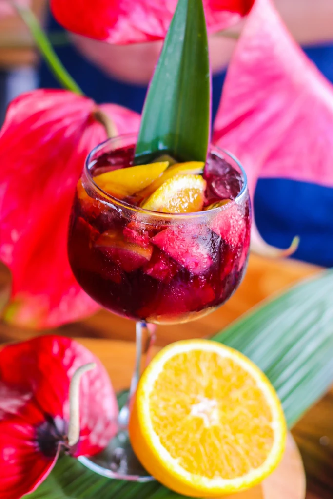 Red fruit drink with ice in wine glass, orange and tropical leaf.