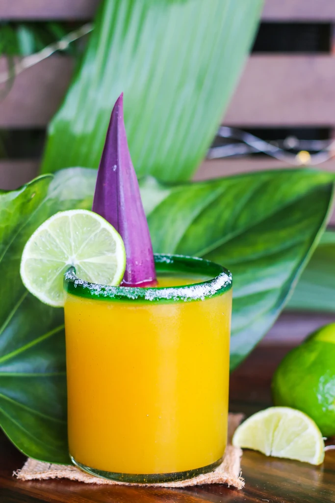 Yellow cocktail with green salted rim, lime, and purple leaf.