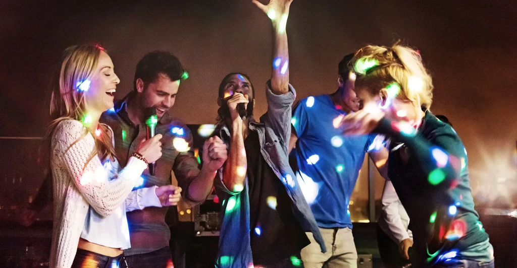 Five people sing and dance enthusiastically at a lively karaoke party.