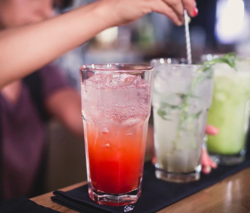 Close-up: Person stirring three vibrant iced drinks on a bar.