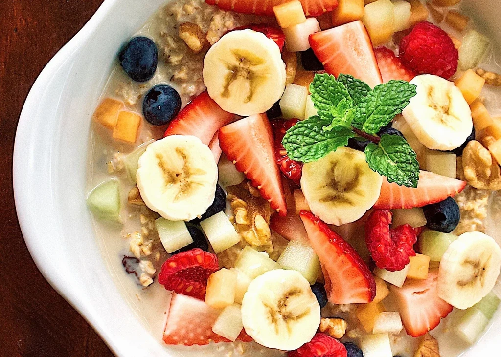 Bowl of oatmeal with fresh berries, banana, nuts, and mint garnish.