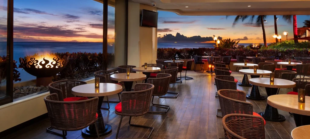 Tropical restaurant patio with glowing tables, ocean sunset, and fire pit.
